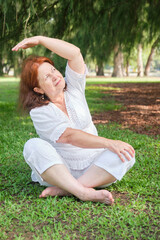 Naklejka na ściany i meble Portrait of a senior Hispanic woman in white clothes practicing yoga outdoors in a park. Concepts: wellness, vitality, active and healthy lifestyle.
