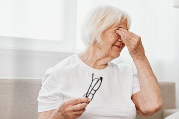 Elderly woman severe eye pain sitting on the couch, health problems in old age, poor quality of...