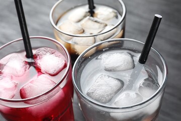 Glasses of different refreshing soda water with ice cubes and straws on grey table, closeup