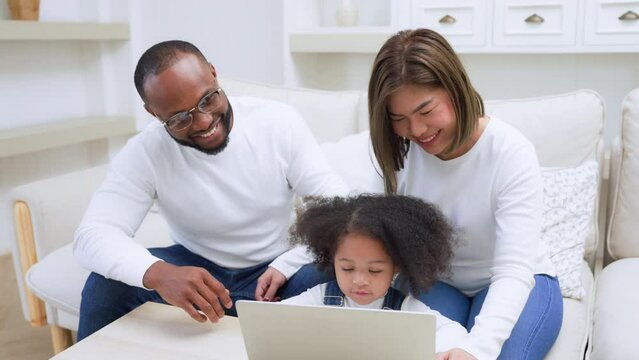 Happy african family parents and little children enjoy using computer laptop together sit on sofa, technology addicted couple with kids having fun at home