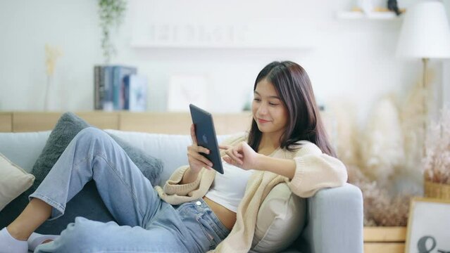 Happy young asian woman relax on comfortable couch at home texting messaging on tablet, smiling girl use tablet, chatting online message, shopping online from home