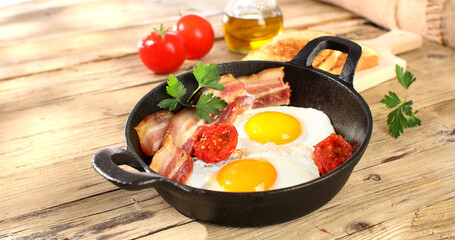 fried eggs with bacon and tomatoes in a frying pan. breakfast on the wooden table. sunny morning. country style. organic farm food