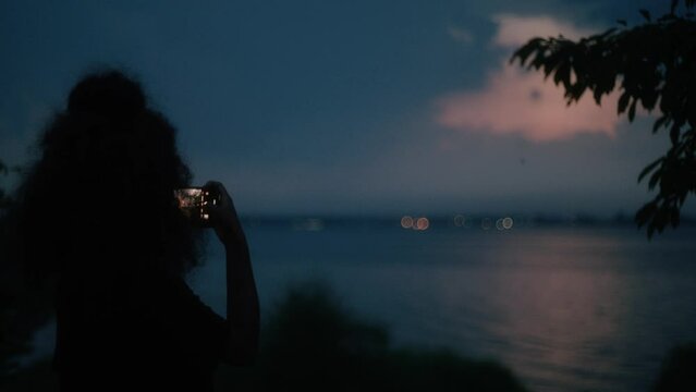 Woman taking photos of sunset on her smartphone, idyllic scenery during dusk time over the water surface.