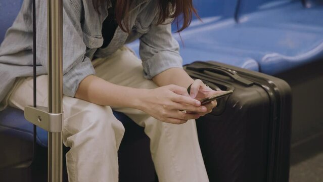 Woman tapping on her smartphone while using public transportation, sitting on blue seat and having a traveling luggage bag near her, luggage and travel accessories concept.