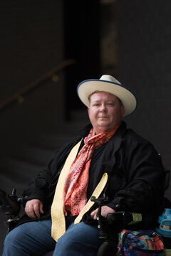 An older LGBTQ person sits in their wheelchair, posing with a confident smile. Their clothing is bright and colorful, expressing their pride in their trans and queer identity.