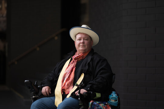 A disabled man smiles confidently at the camera. He is in his powerchair, and sitting outside of a stairwell at an office building. He is trans, and self-identifies as queer.