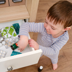 Toddler baby opened the cabinet drawer with pills and medicine. Child boy holding a pack of pills...