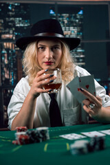attractive girl in shirt and hat drinking whiskey and looking at poker cards in casino - obrazy, fototapety, plakaty