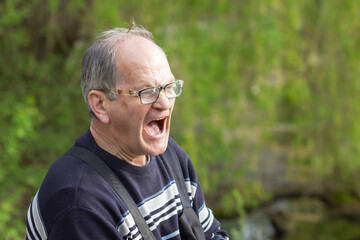 Portrait of screaming or yawning senior man with open mouth in glasses, green nature background....