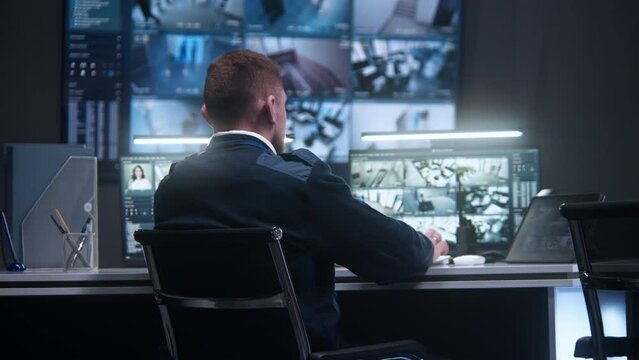 Male officer controls security cameras with AI face scanning. Female operator works in police surveillance center. Big digital screens on the wall and PC monitors with CCTV cameras footage. Back view.