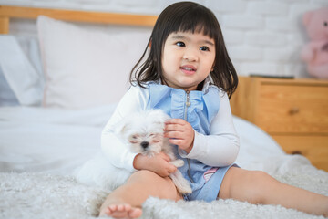 Little girl's sitting and her face lit up with joy as she holding her adorable white dog close on the bed in the cozy bedroom.
