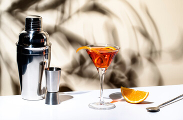 Martinez orange cocktail drink with red vermouth, liqueur, bitter, citrus zest and ice in martini glass. Light beige background, hard light, shadow pattern