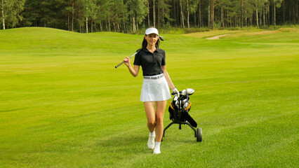 Woman in stylish summer golf outfit walking with bag of drivers on beautiful green course for golf...