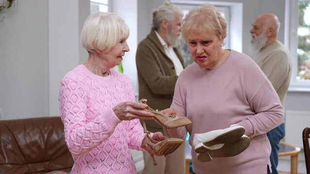 Confident senior women discussing fashion bragging new shoes as blurred men talking at background in living room. Old Caucasian female friends showing off meeting on weekend with husbands