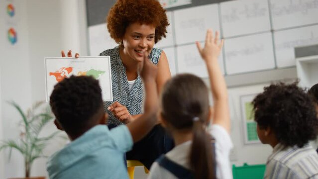 Female elementary school teacher holding map teaching geography class with students raising hands to answer question - shot in slow motion