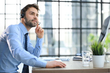 Cheerful young support phone male operator in headset, at workplace while using computer, help service and client consulting call center concept