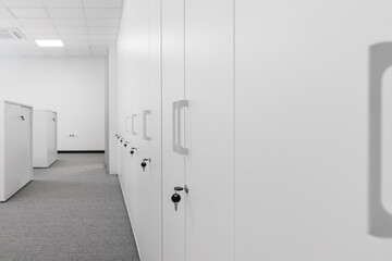 A row of white office cabinets, locked with a key, are located in the aisle of the office space. Shrinking perspective. Selective focus.