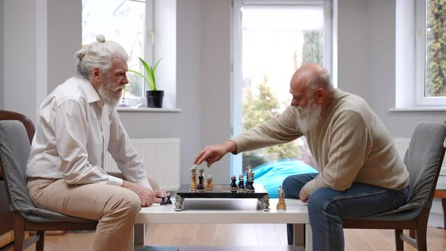 Positive smart senior Caucasian man winning in chess shaking hands with old friend. Side view portrait of happy relaxed retirees enjoying game indoors