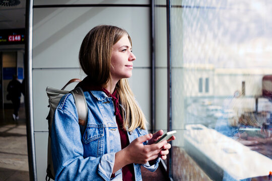 Young woman looking out of window holding smart phone at airport