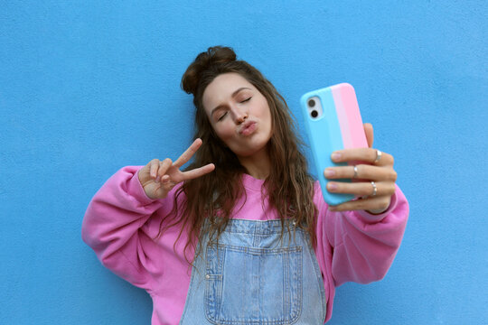 Young woman puckering and taking selfie with peace sign against blue background