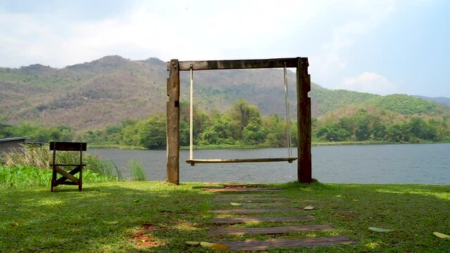 The swing on the verdant green grass field swings back and forth, offering a picturesque view of the tranquil lake, hills, and a gentle breeze blowing by
