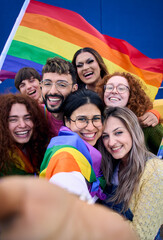 Vertical selfie of LGBT group of young people celebrating gay pride day holding rainbow flag together. Homosexual community smiling taking cheerful self portrait. Lesbian couple friends generation z.