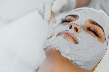 Obraz na płótnie Canvas A woman enjoys a facial treatment in a beauty spa. The beautician puts a mask on the girl's face with a spatula. Camera close-up. The woman lies relaxed, enjoying the procedure