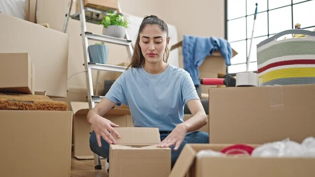Young beautiful hispanic woman packing cardboard box at new home