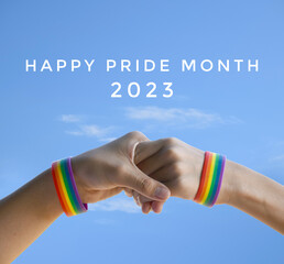 'Happy Pride Month 2023' on bluesky and rainbow wristband in hands background, concept for LGBT people celebrations in pride month, june, around the world.