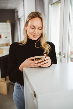 Young blonde woman standing by a cabinet in the office and looking at her phone