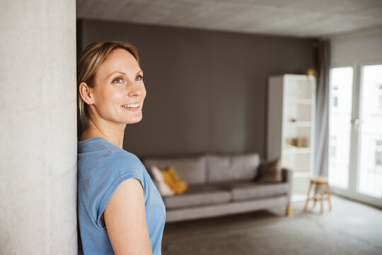 Happy 30-year-old blonde woman in blue t-shirt leaning against a pillar in her apartment and smiling