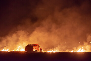 Terrible wild huge fire on the horizon at night in the field. Fire engine on a background of flames and smoke. A large fire scorched the vegetation on the side of the road. - obrazy, fototapety, plakaty