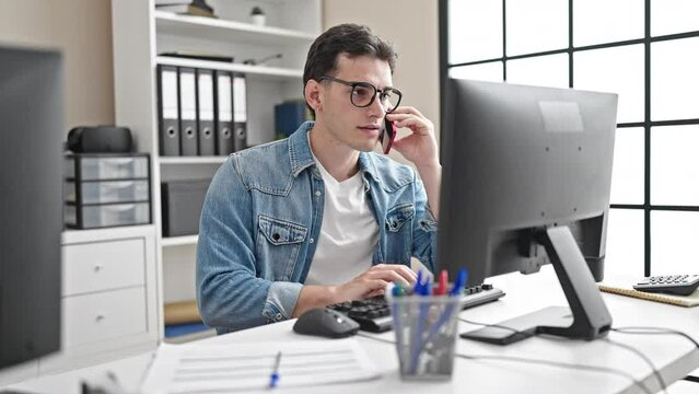 Young hispanic man business worker using computer talking on smartphone at office