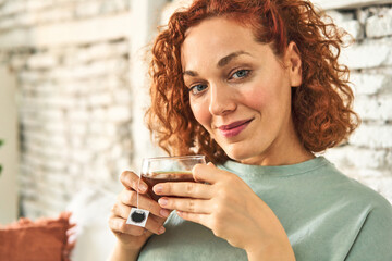 Young, redheaded woman sitting on her home sofa, enjoying a hot cup of tea in natural light.