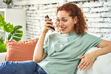 Redhead woman sending voice message on couch, morning light.