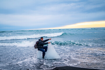 Naklejka premium happy man sitting on big piece of ice on the shore of sea at black beach in iceland