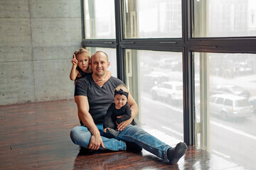A father with two daughters is sitting on the floor by a large window