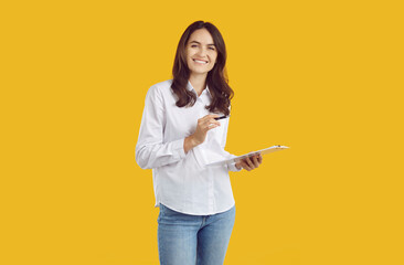 Portrait of happy young female secretary, office manager or business assistant with clipboard. Pretty woman in white shirt standing isolated on yellow background, holding clipboard and pen and smiling - obrazy, fototapety, plakaty