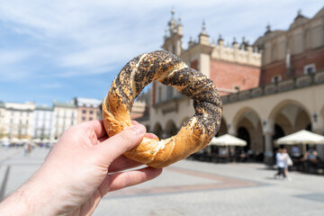 Hand holding obwarzanek krakowski prezel on Kraków Main Market Square, with Cracow Cloth Hall...