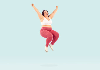 Sporty overweight young woman jumping, celebrating win and triumph. Happy curvy, plus size woman in sportswear rejoicing at her success in losing weight on isolated studio background