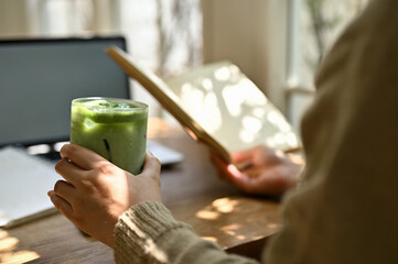 A female sipping iced green tea and reading a book while relaxing at a cafe.