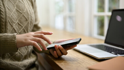 Fototapeta premium A woman in cozy sweater using her smartphone at her workspace.