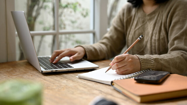 A young Asian woman taking notes on her notebook and using her laptop at a coffee shop.