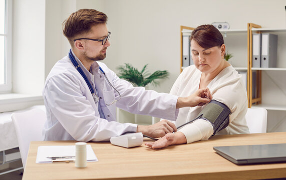 Male doctor working with female patient who has hypertension. Young man physician in white coat sitting at desk with fat, obese woman and using sphygmomanometer to measure her arterial blood pressure