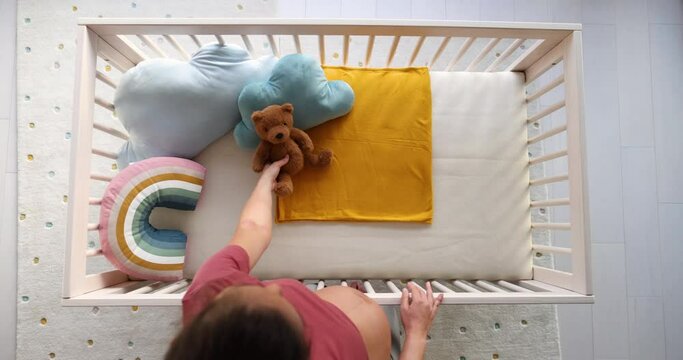 Pregnant woman preparing nursery showing belly baby bump by crib putting teddy bear in crip. Pregnancy concept and home nusery planning. Top view video in slow motion