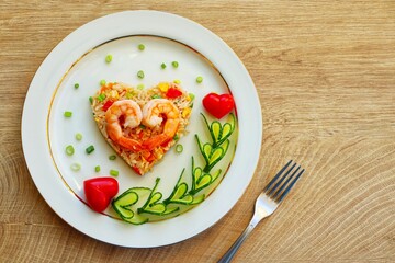 Heart shaped shrimp fried rice served with heart shaped cucumbers and tomatoes on plate with wooden background.Creative art food idea for Valentine or Mother day.Top view.Copy space