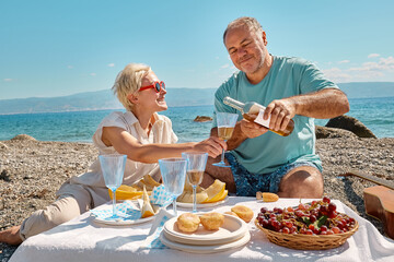 Summer beach picnic. Middle aged couple having picnic with fresh fruit, wine and appetizers outdoors at seaside at the beach. Bearded man pouring wine in wine glass for blond woman.