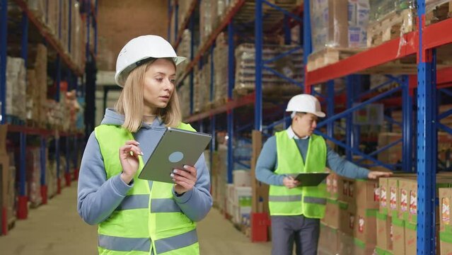 Focused caucasian woman standing among spacious storehouse and typing on digital tablet while asian man writing on clipboard on background. Two coworkers wearing safety helmet and reflective vests.