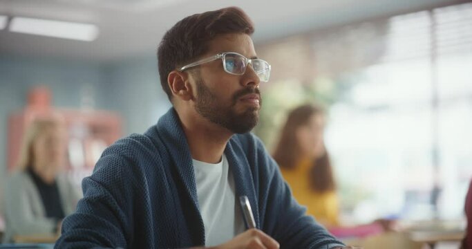 Portrait of an Indian Student Taking a Course in an International Adult Education Center. Young Man Wearing Glasses, Sitting Behind a Desk, Writing Down Notes Together with Diverse Colleagues