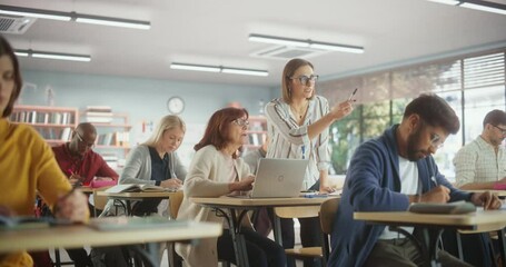 Young Female Teacher Giving a Lecture During an Adult Education Course in School, Having a Conversation with a Older Female with Laptop. Diverse Mature Students Doing Textbook Exercises in Classroom